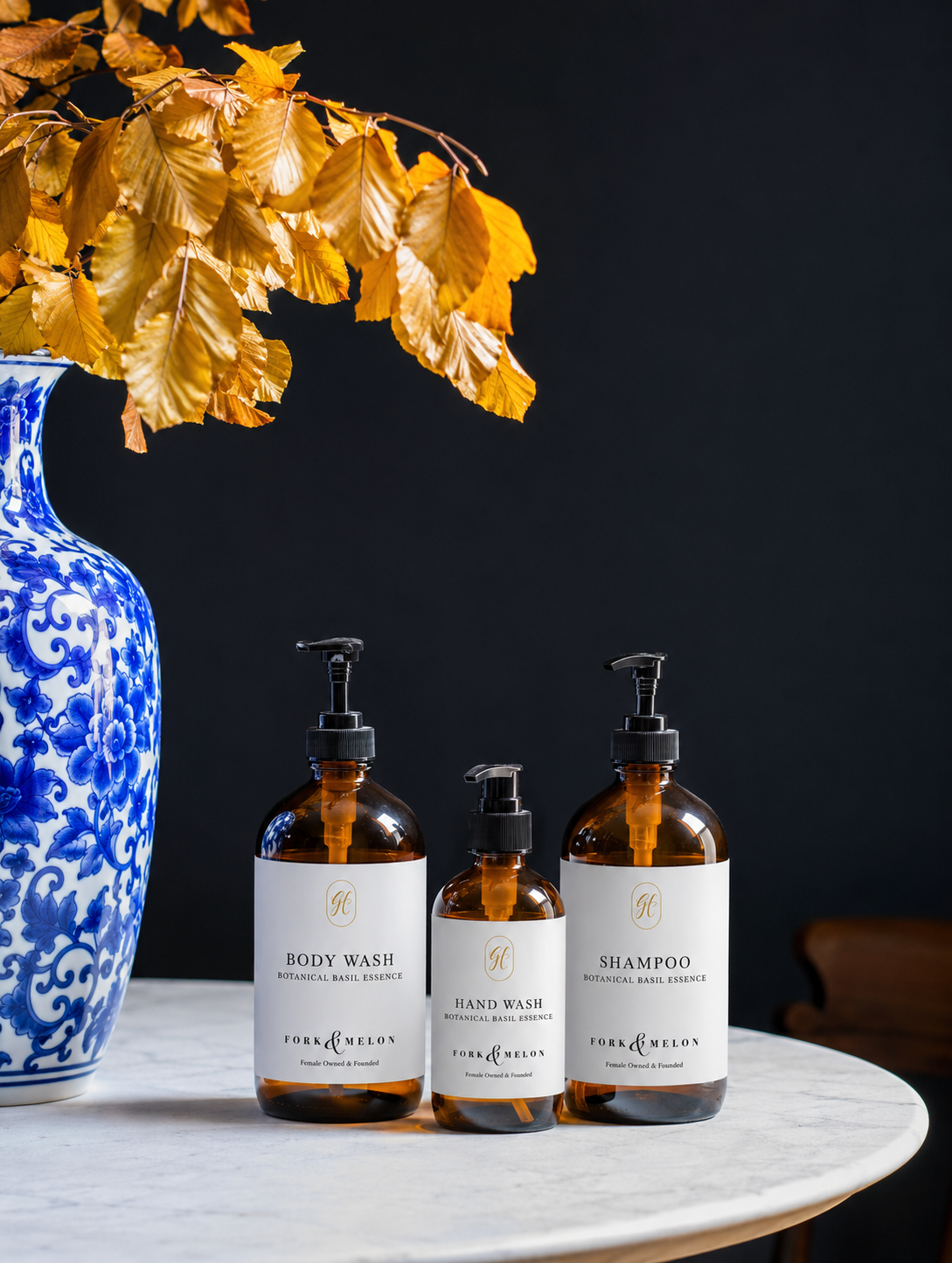 FORK & MELON custom co-branded bottle design created for Guild House Hotel. Three amber pump bottles of personal care products, including body wash, hand wash, and shampoo, arranged on a white marble surface next to a blue and white patterned vase with yellow autumn leaves against a dark background.