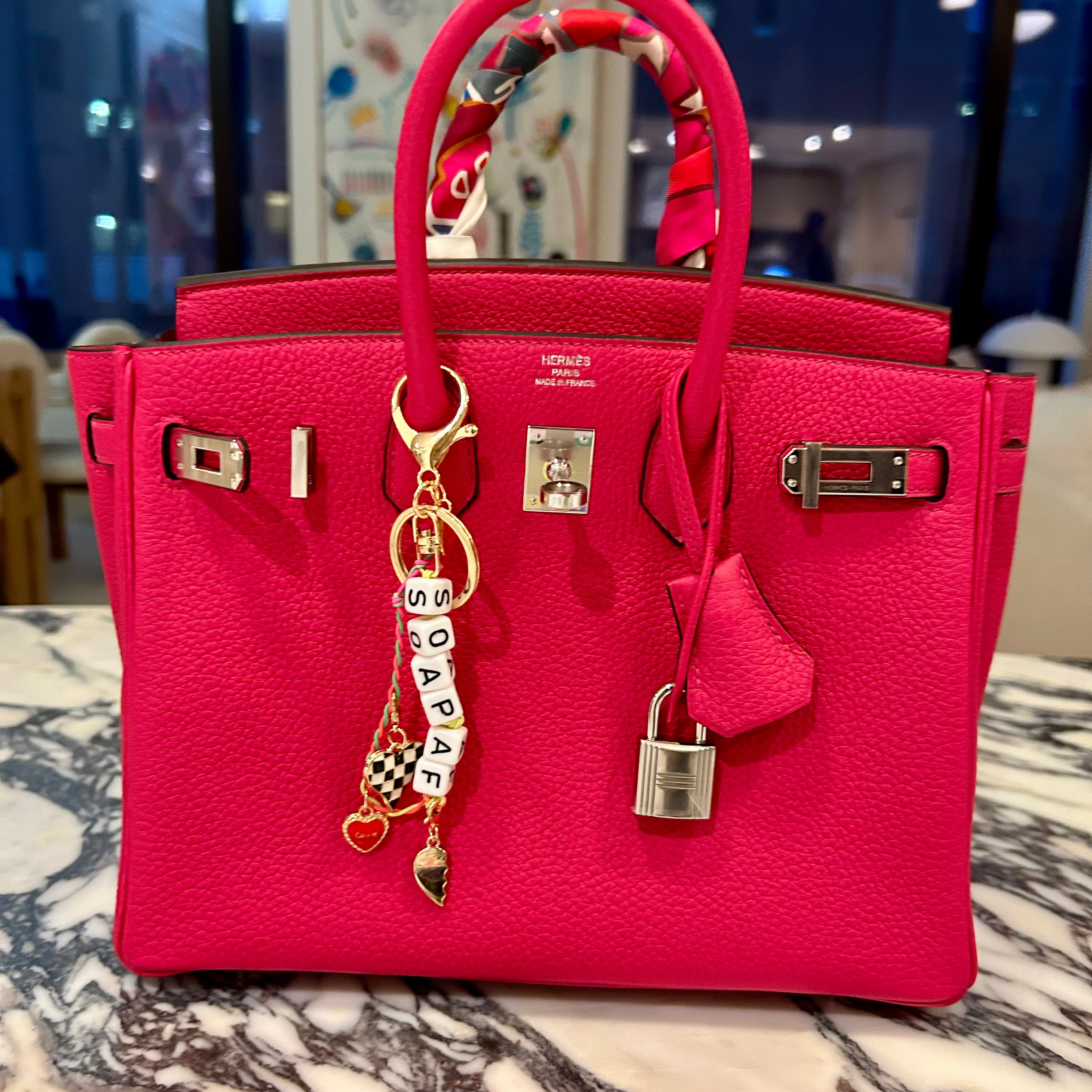 FORK & MELON SOAP AF beaded keychain charm on a bright pink designer Hermes handbag — black and white beaded detail with gold hardware and checkered heart accent, styled as a chic accessory.